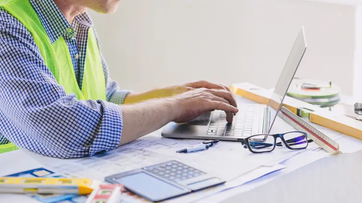 Person in a hi-vis vest typing on a laptop beside a calculator and drawings