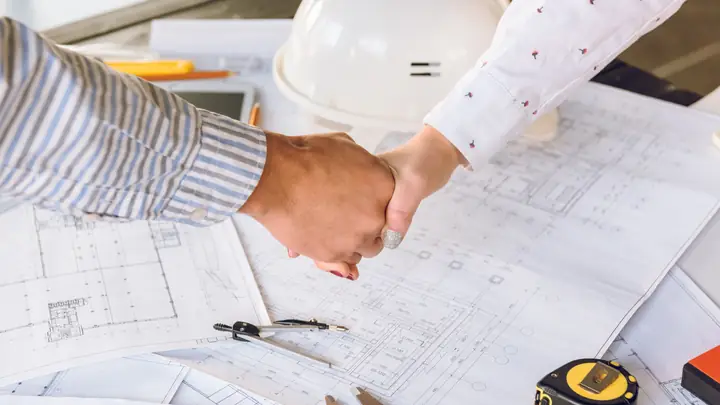 Handshake over drawings with a white hard hat and tape measure