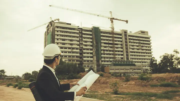 Man in a suit and hard hat holding drawings in front of a large building with cranes