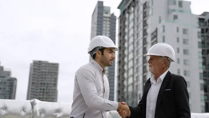 Two men in hard hats shaking hands with tower blocks behind