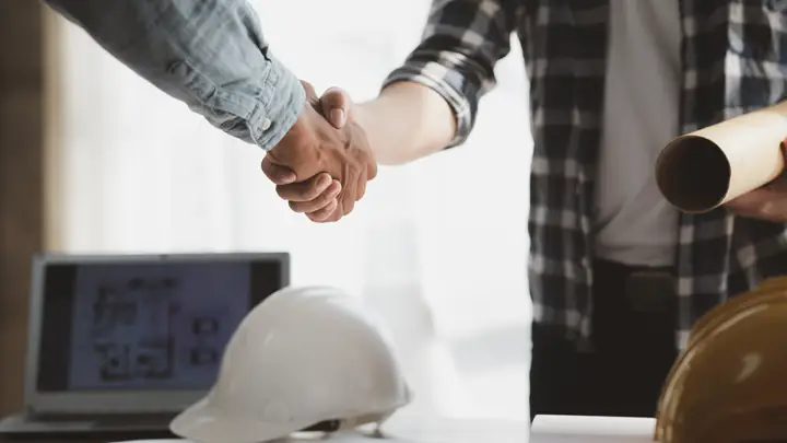 Handshake by a desk with a laptop showing plans and a white hard hat