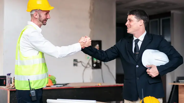 Site manager in a hi-vis vest shaking hands with a man in a suit holding a hard hat