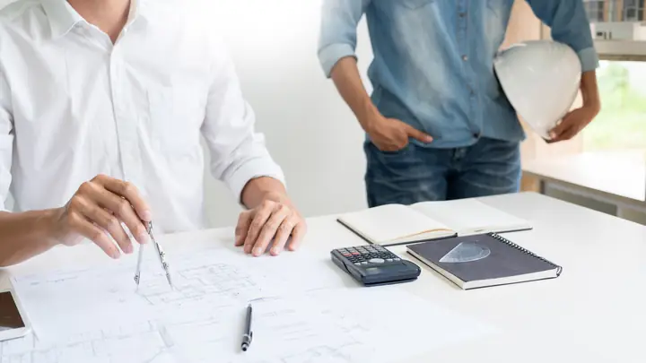 Two people at a desk with drawings, a calculator and a hard hat