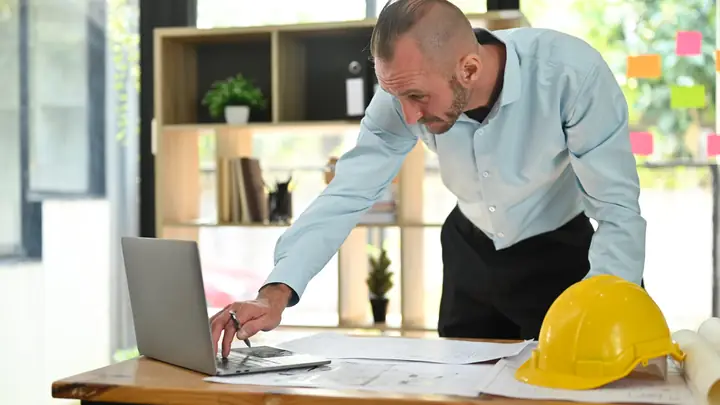 Man leaning over a laptop and drawings at a desk with a yellow hard hat