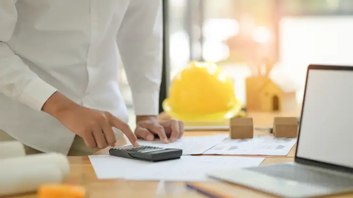 Hands using a calculator next to a laptop, drawings and a yellow hard hat