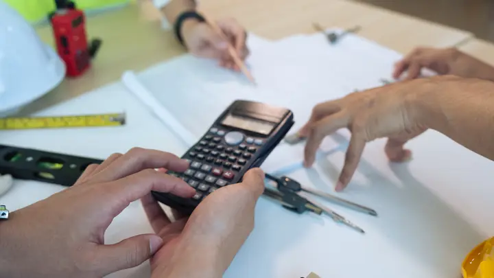 Hands using a calculator over drawings with a level and dividers
