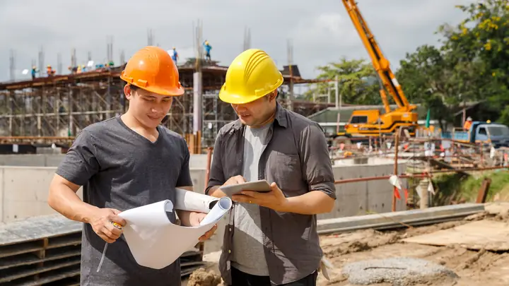 Two men in hard hats with drawings and a tablet on a busy site