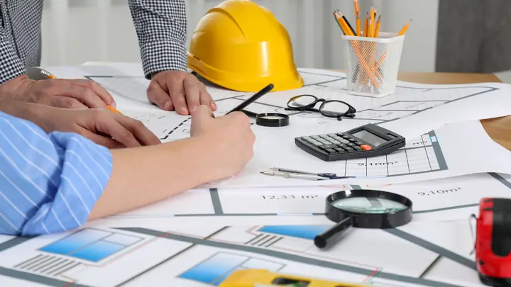 Hands over drawings with a calculator, magnifying glass and yellow hard hat