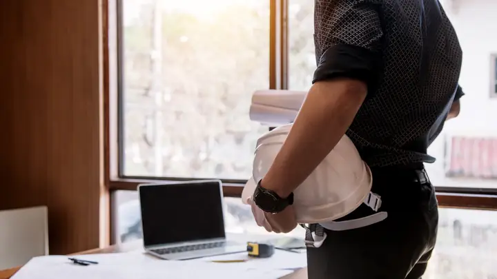 Man holding a white hard hat and rolled drawings by a desk with a laptop