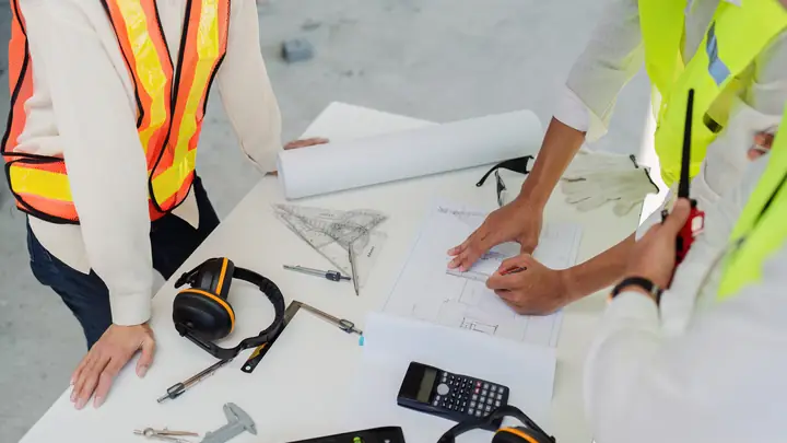 Two people in hi-vis vests working over drawings with a calculator and ear defenders