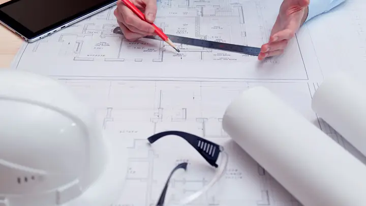 Hand drawing on plans with a ruler, white hard hat and safety glasses nearby