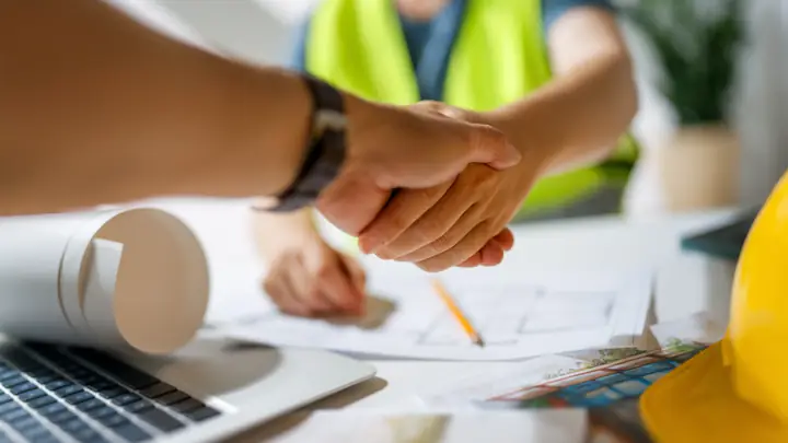 Handshake over a laptop and drawings, colleague in hi-vis behind
